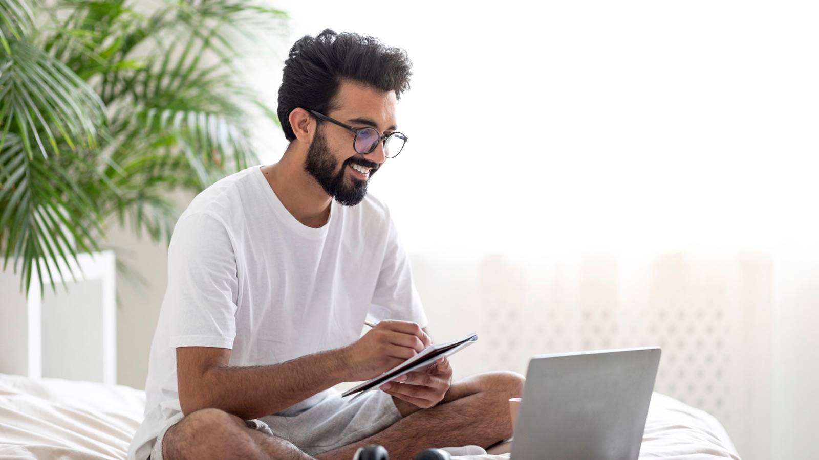 Man writing in a notepad on his bed beside a laptop